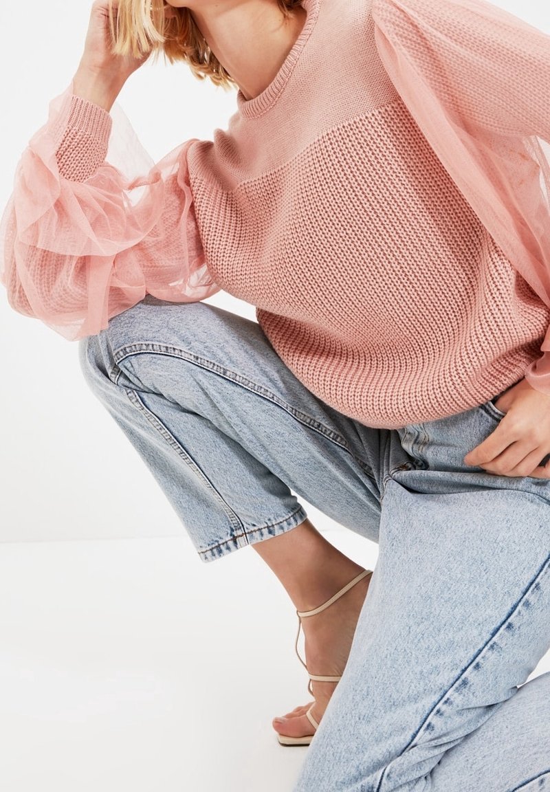 Rosa stickad tröja med genomskinliga, volangärmar, matchad med ljusblå denimjeans. Sandaler har tunna remmar.