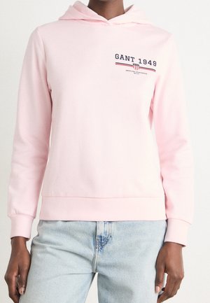 Persoon in een lichtroze capuchontrui met de tekst "GANT 1949" en lichtblauwe spijkerbroek, getoond van nek tot heupen.