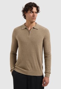 Beige lange mouwen pullover in een gestructureerde brei met een ritssluiting bij de kraag, met een aansluitend ontwerp en geribbelde manchetten en zoom. Draagt met zwarte broek.