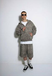 Szary, wzorzysty hoodie z zamkiem i dopasowane szorty, w połączeniu z białą koszulką, czarnymi trampkami i okularami przeciwsłonecznymi. Gładka faktura.