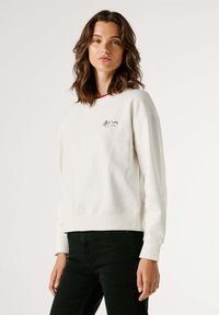 Biela mikina s okrúhlym výstrihom a červeným lemom na golieri, s malým čiernym vyšívaným motívom na hrudi. Mäkká textúra a uvoľnený strih.