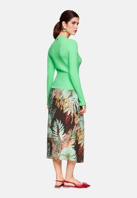 Grünes strukturiertes Langarmoberteil kombiniert mit einem floral gemusterten braunen Rock, der tropische Blätter zeigt, ergänzt durch rote Slingback-Schuhe.