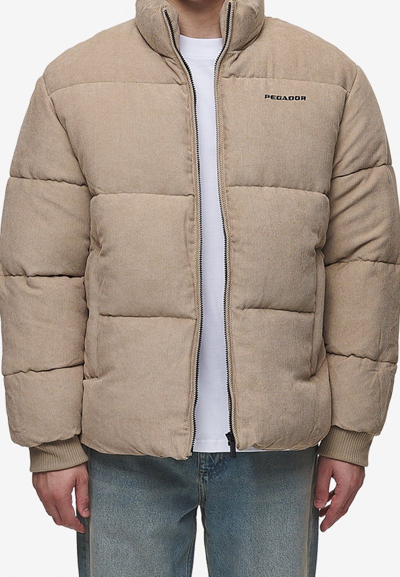 Veste matelassée en velours côtelé beige avec fermeture éclair portée sur une chemise blanche et un jean bleu clair, arborant le logo "PEGADOR" sur la poitrine.