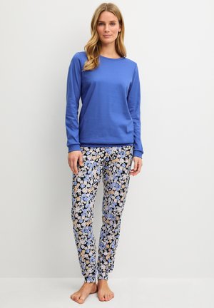 Vrouw die een blauw shirt met lange mouwen en een broek met bloemenprint draagt, staat blootsvoets tegen een eenvoudige lichte achtergrond.