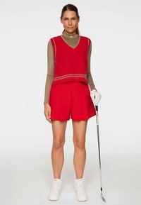 Gilet rouge en maille porté sur un haut à manches longues beige, assorti à un short plissé rouge. Porte des baskets blanches et tient un club de golf.