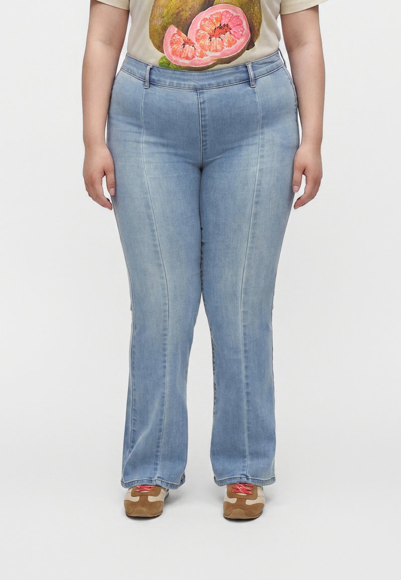 Person iført lyseblå bootcut jeans, beige t-shirt med frugtprint og brune sneakers med lyserøde snørebånd, stående på hvid baggrund.