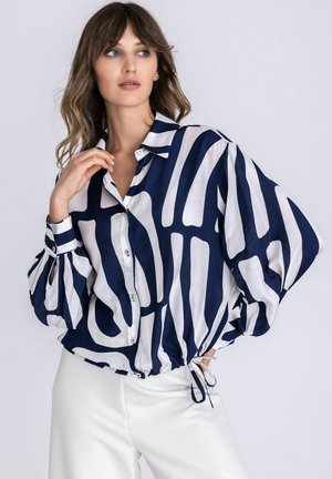 Femme portant un chemisier à manches longues boutonné avec des rayures abstraites bleu marine et blanches, associé à un pantalon blanc, posant devant un fond uni.