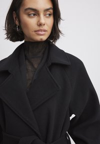 Abrigo de mezcla de lana negra con un amplio collar y diseño envolvente. Cuenta con una cintura ceñida con cinturón y una blusa negra en capas debajo con un acabado texturizado.
