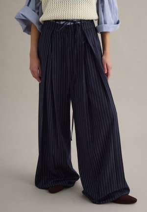 Person trägt marineblaue, weit geschnittene Nadelstreifenhose mit einer cremefarbenen Strickweste über einem blau gestreiften Hemd mit hochgekrempelten Ärmeln.