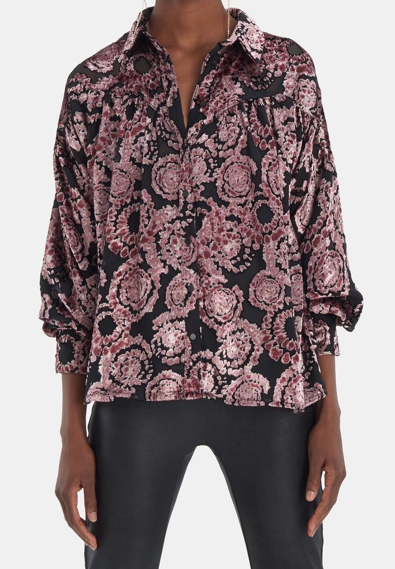 Femme portant un chemisier noir ample avec des motifs floraux circulaires roses en velours et un pantalon noir ajusté, debout, les bras relâchés le long du corps.
