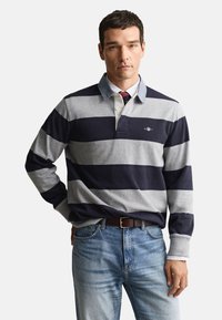 Polo de manga comprida com riscas azul-marinho e cinza, com gola e pala com botões. Usado com jeans azul claro e cinto castanho.