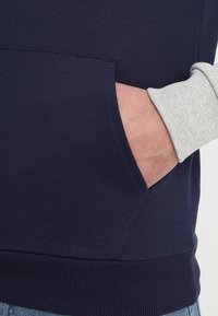 Sweatshirt bleu marine avec un ourlet côtelé et des manches longues gris clair. Présente une poche sur le devant, une texture de tissu lisse et un design simple.