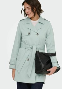 Femme portant un trench-coat vert clair ceinturé, tenant un sac à main en cuir noir, regardant vers le bas, sur un fond clair uni.