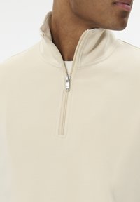 Poloppulóver so zipsom do polovice v béžovej látke. Má stojačikový golier a hladkú textúru so strieborným detailom zipsu na krku.