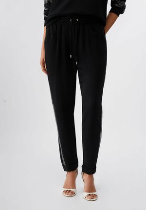 Donna che indossa pantaloni neri a sigaretta con vita a coulisse e strisce laterali di strass, abbinati a tacchi bianchi aperti.