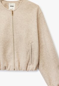 Beige kort jakke lavet af tekstureret stof, med rund halsudskæring, frontlynlås og sidelommer. Accentueret med en subtil glans.