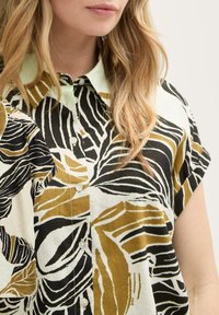 Blusa a maniche corte con stampa di foglie bianca e nera con accenti oliva, colletto verde chiaro e chiusure a bottone lungo il fronte.