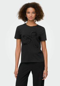 Kvinde med krøllet hår iført en sort kortærmet top med et tekstureret bånd-design og matchende sorte bukser, stående foran en hvid baggrund.