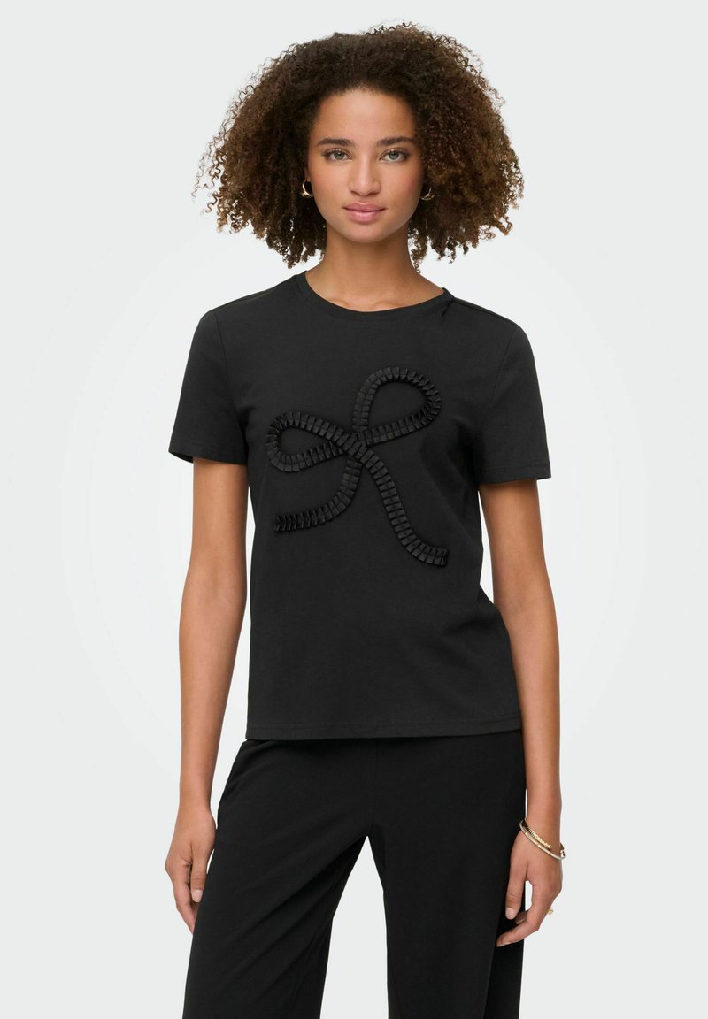 Kvinde med krøllet hår iført en sort kortærmet top med et tekstureret bånd-design og matchende sorte bukser, stående foran en hvid baggrund.