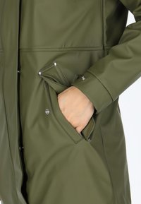 Giacca impermeabile verde oliva con una texture liscia, design aderente e dettagli con bottoni a pressione. Presenta una tasca laterale con un cappuccio pieghevole.