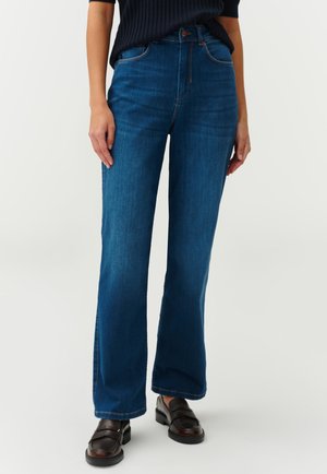 Blue denim flared jeans featuring a high waist, smooth texture, contrast stitching, and a subtle faded wash, paired with black loafers.