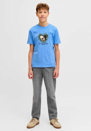 Adolescent garçon aux cheveux bouclés portant un t-shirt bleu avec un motif de crâne, un jean gris et des chaussures beiges à lacets, debout devant un fond uni.