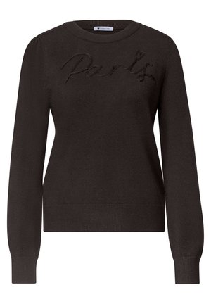 Donna che indossa un maglione nero con testo "Paris", jeans blu, sneakers bordeaux e una borsa a tracolla marrone, in piedi contro un muro bianco sorridendo.