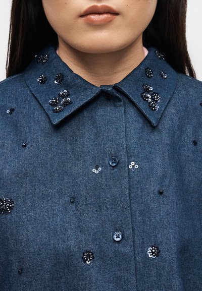 Femme portant une chemise en denim bleu avec des boutons sur le devant et des embellissements de fleurs en sequins noirs et argentés sur le col et le tissu.