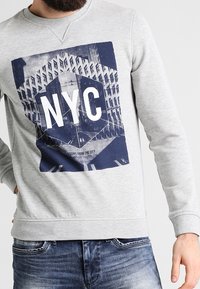Sudadera gris con una gran impresión gráfica de NYC con letras blancas en negrita. El diseño incluye imágenes arquitectónicas y un borde hexagonal.
