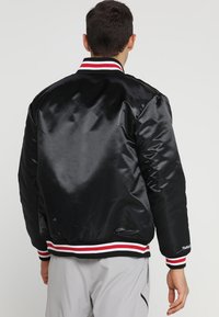 Sort satin bomberjakke med ribbetkrave og -manchetter med røde og hvide striber. Kviltet struktur uden synlig branding på ryggen.