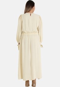 Long, cream-colored dress with a keyhole back, elastic waist, pleated shoulders, and tie accents on the sleeves. Smooth, lightweight fabric.