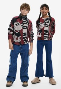 Twee kinderen dragen gebreide truien met opvallende Mickey Mouse graphics in rood en zwart, gecombineerd met blauwe flared jeans en casual schoenen.