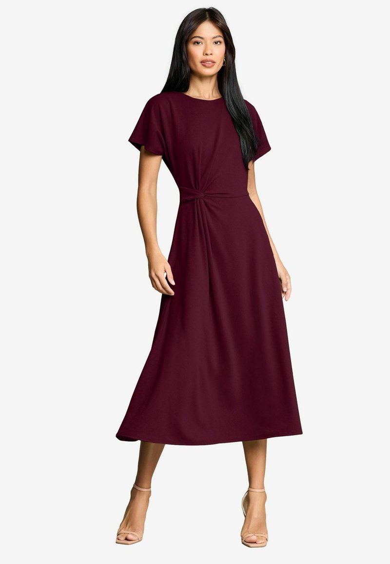 Burgundy midilång klänning med korta ärmar, med en knuten midjedetalj och mjukt, flödande tyg. Bärs med nude högklackade sandaler.