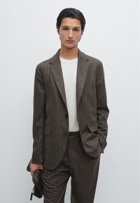Braune Nadelstreifen-Sakko mit Revers, einem Einzelknopf und Vorderaschen, kombiniert mit einem hellbeigen Pullover und hält eine dunkelbraune Tasche.