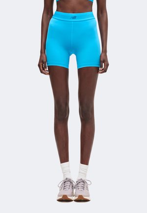 Person iført lyseblå træningsshorts, hvide sokker og grå løbesko, der står mod en ensfarvet hvid baggrund.