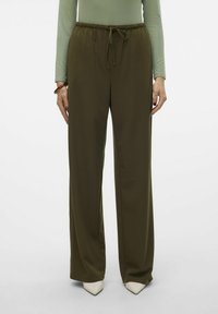 Pantalons larges en vert olive avec une taille élastique et un cordon de serrage. Tissu doux avec une texture lisse ; associés à des chaussures à bout pointu crème.