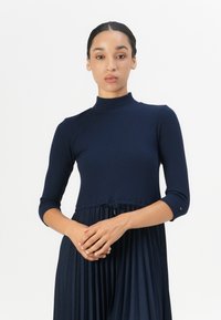 Navy ribbed dress with high neckline, three-quarter sleeves, and pleated skirt. Features a drawstring waist and contrasting accents.