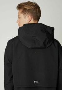 Čierna nepremokavá bunda s kapucňou, s cropped strihom na zadnej strane a bielou grafikou hory na dolnom okraji. Hladká textúra.