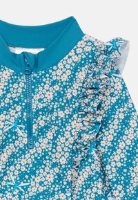 Teal kopalna majica z cvetličnim vzorcem v beli in oranžni barvi, z zadrgo spredaj in nabori na ramenu.