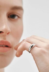 Anillo de metal plateado con un acabado suave y pulido y un diseño curvado. Llevado en una mano, resaltando su superficie reflectante y su forma elegante.