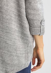 Szary sweter z dzianiny o teksturowanej tkaninie, z podwiniętymi mankietami z ozdobnym guzikiem i luźnym krojem. Widoczny szew na dole.