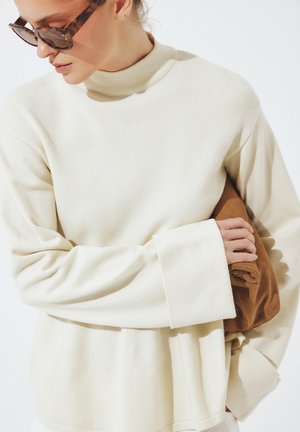 Kobieta w dużym kremowym swetrze i okularach przeciwsłonecznych w oprawkach w kolorze żółwia, trzymająca złożony brązowy ubiór pod pachą.