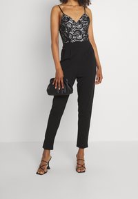 Svart jumpsuit med en spetstop med V-ringning, slim-fit byxor och en svart kuvertväska. Outfiten kompletteras med svarta sandalskor med remmar.