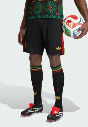 Calciatore maschio dalla vita in giù che tiene in mano un pallone Adidas bianco, indossa pantaloncini neri, calzini neri con strisce rosse, verdi e gialle, e scarpe da calcio.