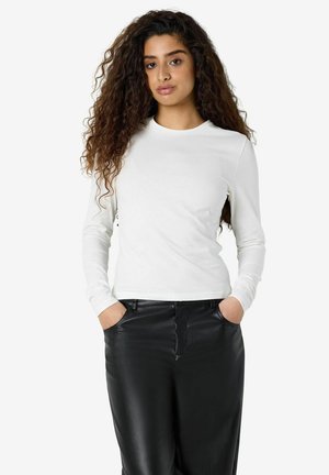 Femme aux longs cheveux bouclés portant une chemise blanche à manches longues et un pantalon en cuir noir, les mains dans les poches, debout devant un fond blanc.