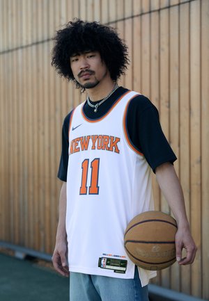 Hvid basketballtrøje med orange detaljer, med "NEW YORK" og nummer "11." Sort undertrøje, blå jeans og holder en brun Spalding basketball.