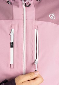 Rosa wasserdichte Jacke mit durchgehendem Reißverschluss, zwei Fronttaschen mit weißen Reißverschlüssen und Kapuze mit Kordelzug. Sie hat eine glatte Textur.