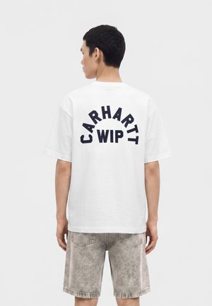 Uomo che indossa una T-shirt bianca a maniche corte con "CARHARTT WIP" in lettere maiuscole sul retro e shorts di denim grigio sbiadito.