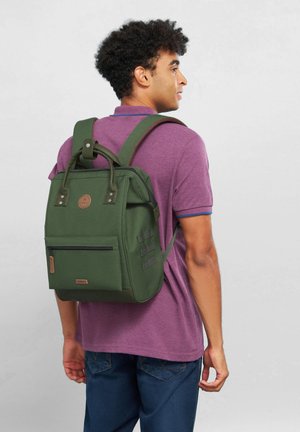 Grüner Rucksack mit strukturiertem Design, oberem Griff, verstellbaren Tragegurten, Fronttasche mit Reißverschluss und strukturierten Akzenten.