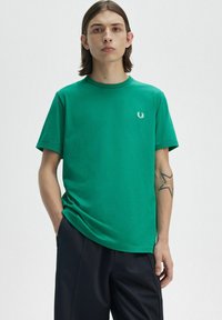 Giovane con capelli lunghi fino alle spalle, indossa una maglietta verde a maniche corte con un piccolo logo bianco, pantaloni neri e un tatuaggio a stella sul lato sinistro dell'avambraccio.